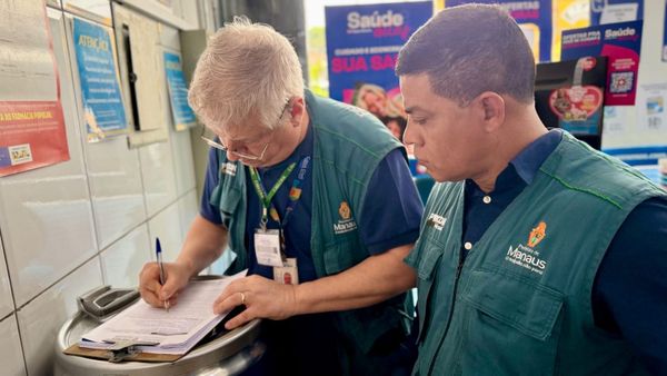 Dois fiscais do Procon Manaus, usando coletes verdes da prefeitura, preenchem formulário durante fiscalização em farmácia.