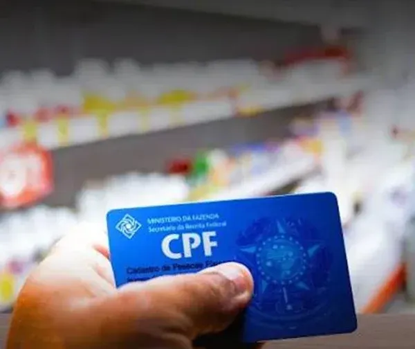 Mão segurando um cartão azul do CPF em frente a prateleiras de medicamentos em uma farmácia.