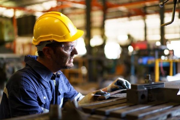 Trabalhador com capacete amarelo e óculos de proteção opera maquinário em uma linha de produção industrial.