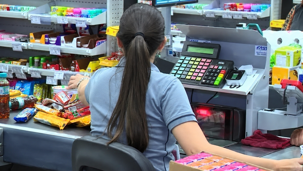 Operadora de caixa de costas trabalhando em um supermercado, registrando produtos em frente a um monitor e teclado.