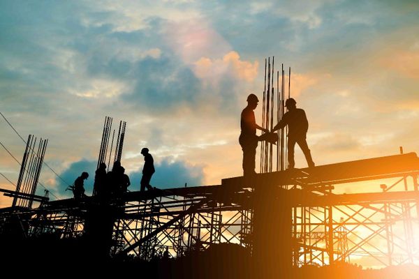 Silhueta de trabalhadores em canteiro de obras de infraestrutura sob o pôr do sol, com vigas de aço e andaimes expostos.