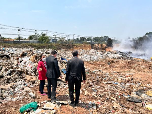 Três pessoas de costas observam área de descarte de resíduos com fumaça ao fundo no Distrito Industrial II, em Manaus.