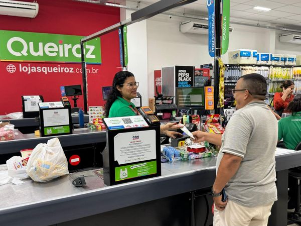 Operadora de caixa atende cliente em unidade das Lojas Queiroz em Manaus, com logomarca da rede ao fundo.