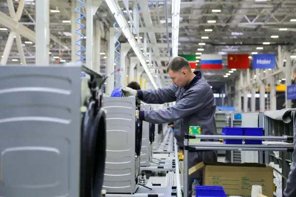 Trabalhador em linha de montagem de máquinas de lavar da Haier, com bandeiras da China e Rússia ao fundo.