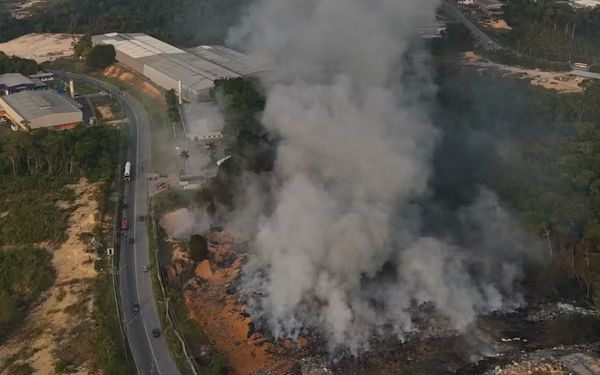 Grande coluna de fumaça branca saindo de área de lixão ao lado de rodovia e galpões industriais em Manaus.