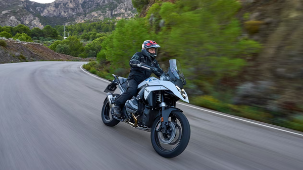 Motociclista em uma moto branca de alta cilindrada percorrendo uma estrada sinuosa cercada por vegetação e montanhas.