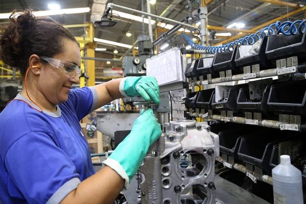Trabalhadora com luvas verdes e óculos de proteção manipula peça metálica em linha de montagem industrial.