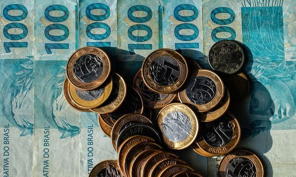 Cédulas de cem reais dispostas verticalmente com diversas moedas de um real espalhadas por cima.