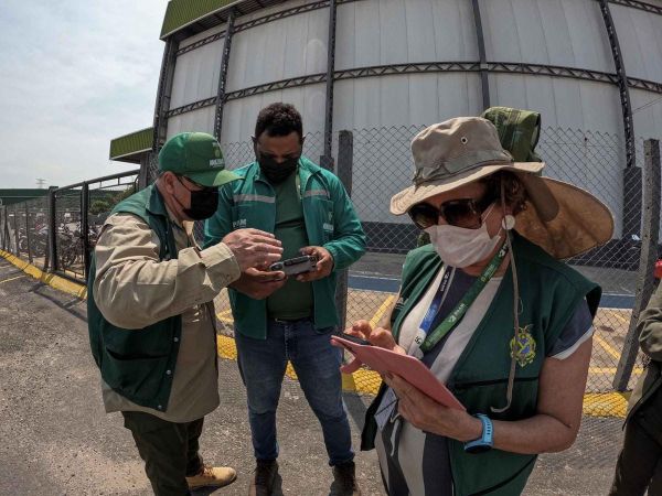 Três fiscais do Ipaam usando coletes verdes e máscaras operam drone e tablet durante vistoria em área industrial de Manaus.