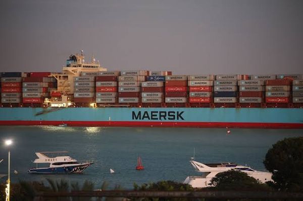 Navio porta-contêineres azul da Maersk carregado navegando em um rio ao entardecer, com lanchas menores em primeiro plano.