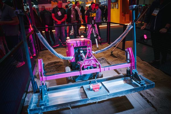 Robô cor-de-rosa de nivelamento de concreto em exposição em um estande, com pessoas ao fundo observando a tecnologia.