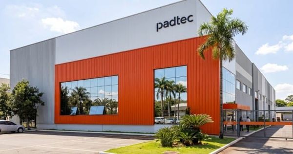 Fachada de prédio industrial da Padtec com detalhes em laranja e cinza, refletindo palmeiras em suas janelas de vidro.