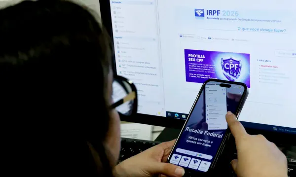 Pessoa segura celular com aplicativo da Receita Federal aberto em frente a monitor com o programa do IRPF 2026.