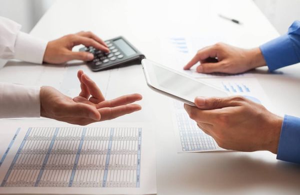 Duas pessoas em uma mesa com calculadora, tablet e papéis com tabelas financeiras, simulando uma negociação de dívidas.