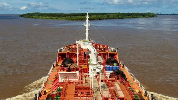 Vista frontal de um navio cargueiro navegando pelas águas barrentas do Rio Amazonas em direção a uma ilha de vegetação.