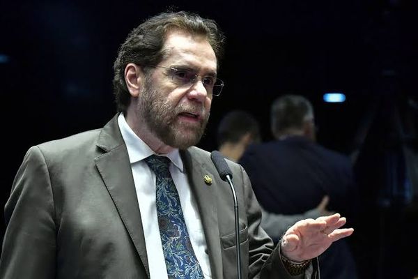 O senador Plínio Valério discursa em um microfone, vestindo terno marrom e gravata estampada.
