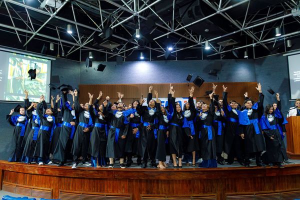 Estudantes de beca preta e azul jogam capelos para o alto em comemoração no auditório da Escola Senai Waldemiro Lustoza.