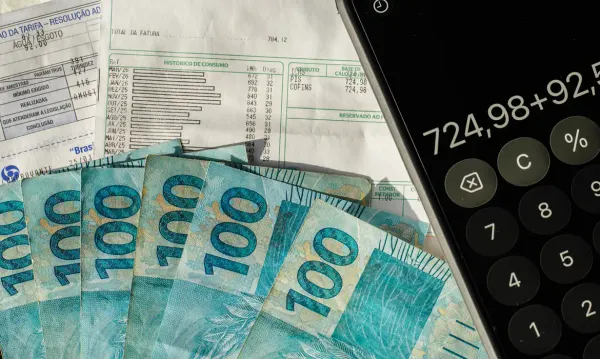 Cédulas de cem reais sobre faturas de consumo ao lado de um smartphone exibindo uma calculadora.