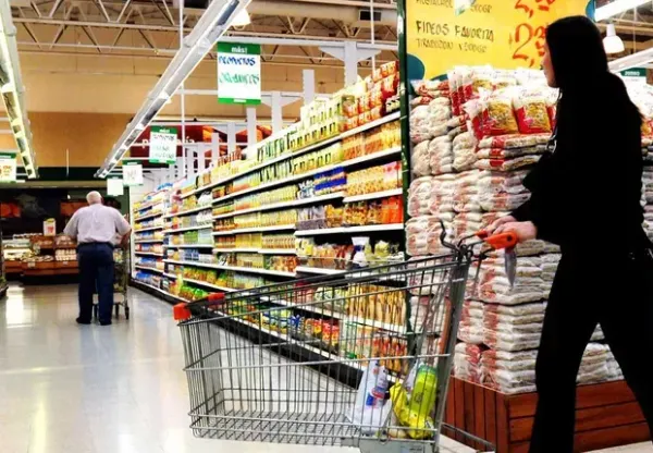 Mulher empurra carrinho de compras em corredor de supermercado com prateleiras abastecidas.