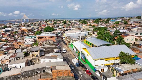 Vista aérea de área urbana densa em Manaus com casas, prédios baixos e a Ponte Rio Negro ao fundo sob céu com nuvens.