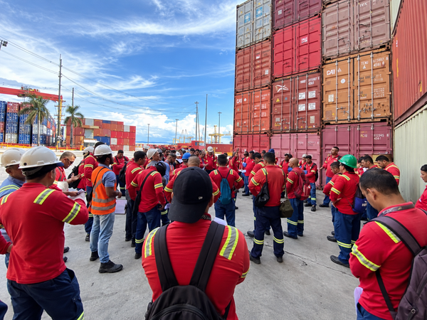 Trabalhadores com uniformes vermelhos e capacetes reunidos em pátio logístico com contêineres empilhados ao fundo.