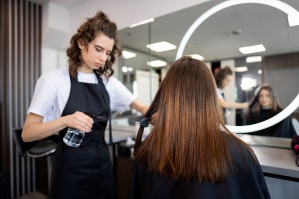 Cabeleireira com avental preto aplica spray no cabelo de uma cliente em um salão de beleza com espelho circular iluminado.