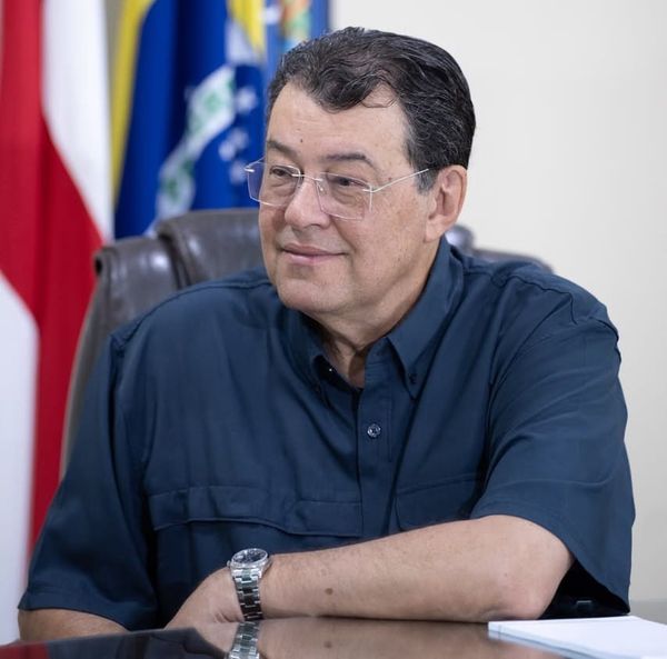 O senador Eduardo Braga, de óculos e camisa azul escura, sentado à mesa com bandeiras ao fundo.