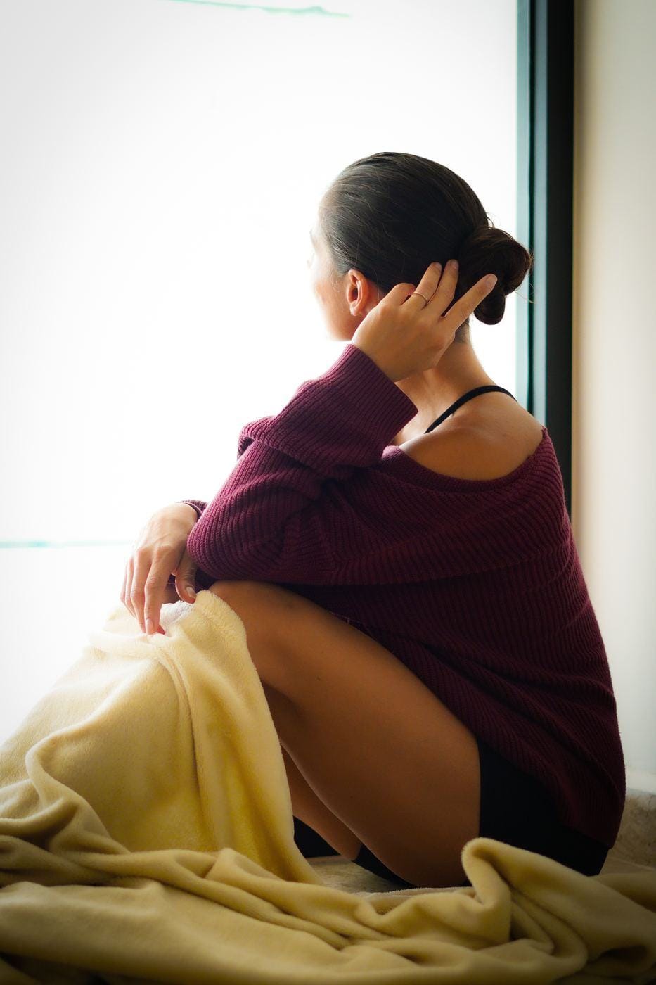 A woman wrapped in a sweater and blanket by a window, at ease, looking outside