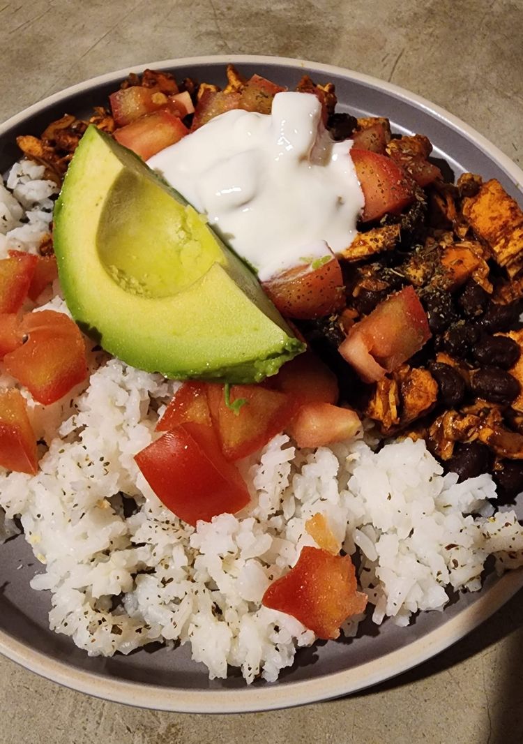 Overhead view of a bowl with white rice, black beans, seasoned chicken, diced tomato, avocado, and Greek yogurt on a concrete surface.