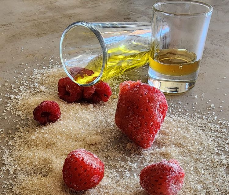 A tipped glass of olive oil and a small glass of honey set among sugar, fresh raspberries, and a strawberry.
