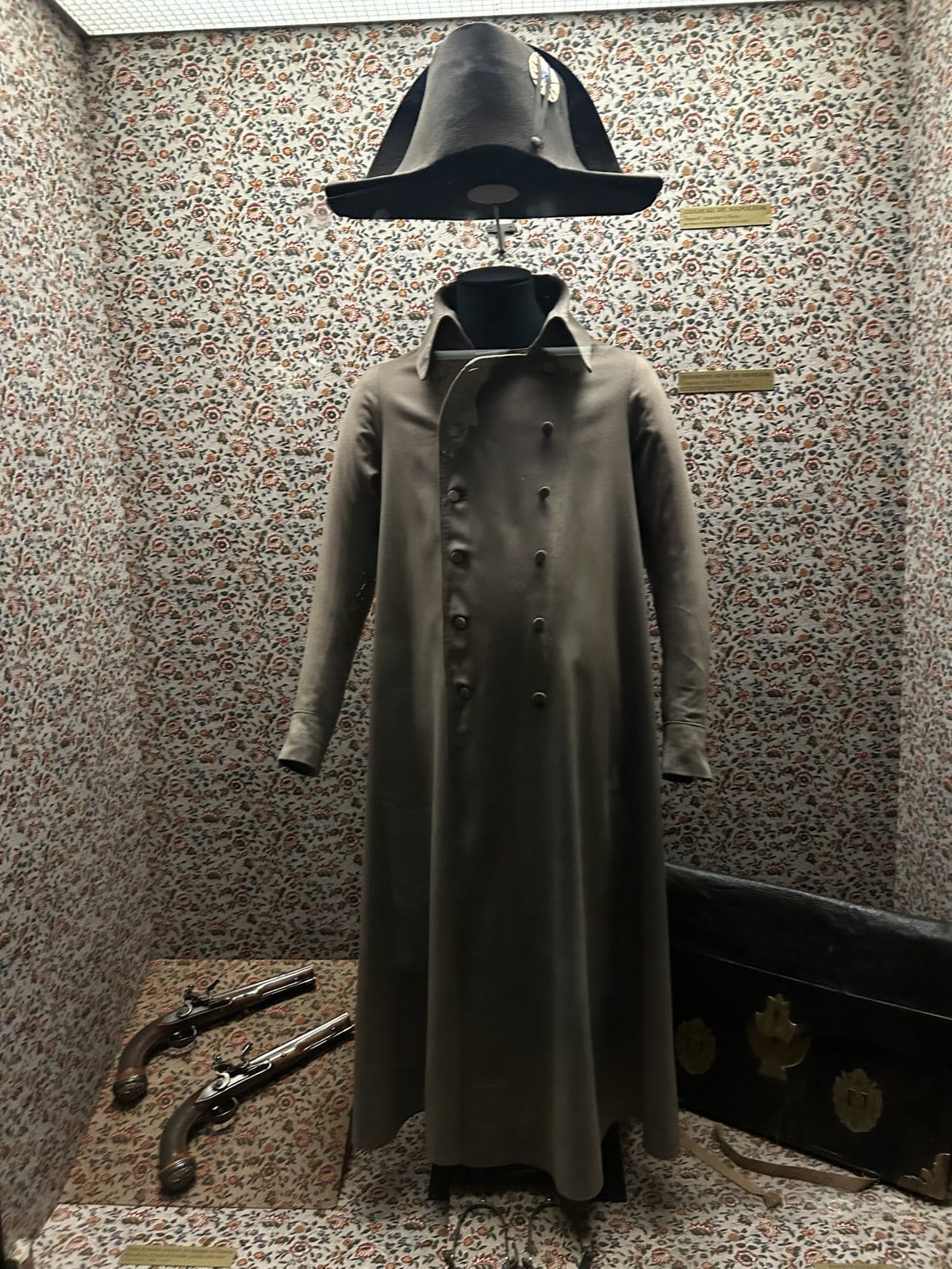 A grey coat and a bicorne hat displayed in a glass case with two pistols