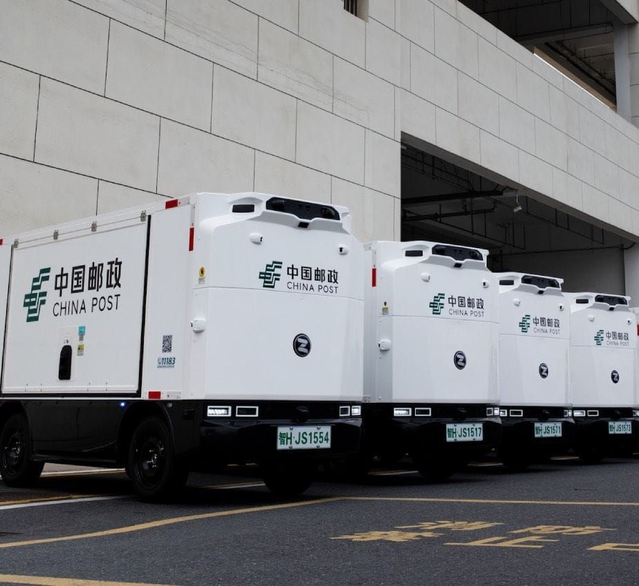 A row of white Zelostech driverless delivery vans in China Post livery parked beside a building.