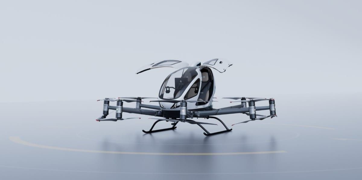 EHang's EH216-S pilotless passenger eVTOL aircraft, white and grey with rotor arms spread, on a plain pale background.