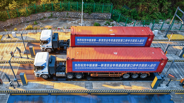 Two driverless container trucks carrying red containers pass through a checkpoint on a freight channel, seen from above.