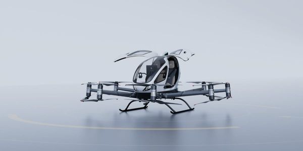EHang's EH216-S pilotless passenger eVTOL aircraft, white and grey with rotor arms spread, on a plain pale background.