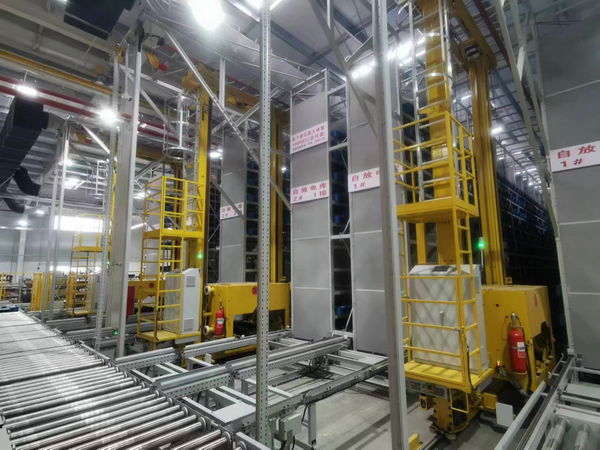 Yellow stacker cranes on rails between tall storage racks in a warehouse, with roller conveyors in the foreground