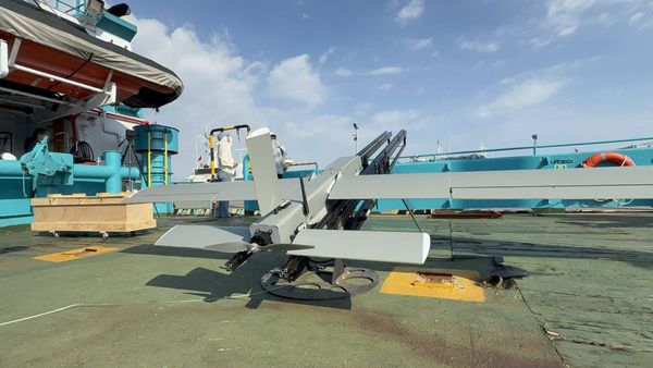 A gray fixed-wing drone on a catapult launch rail on a ship deck under a blue sky, with crew members in the background.