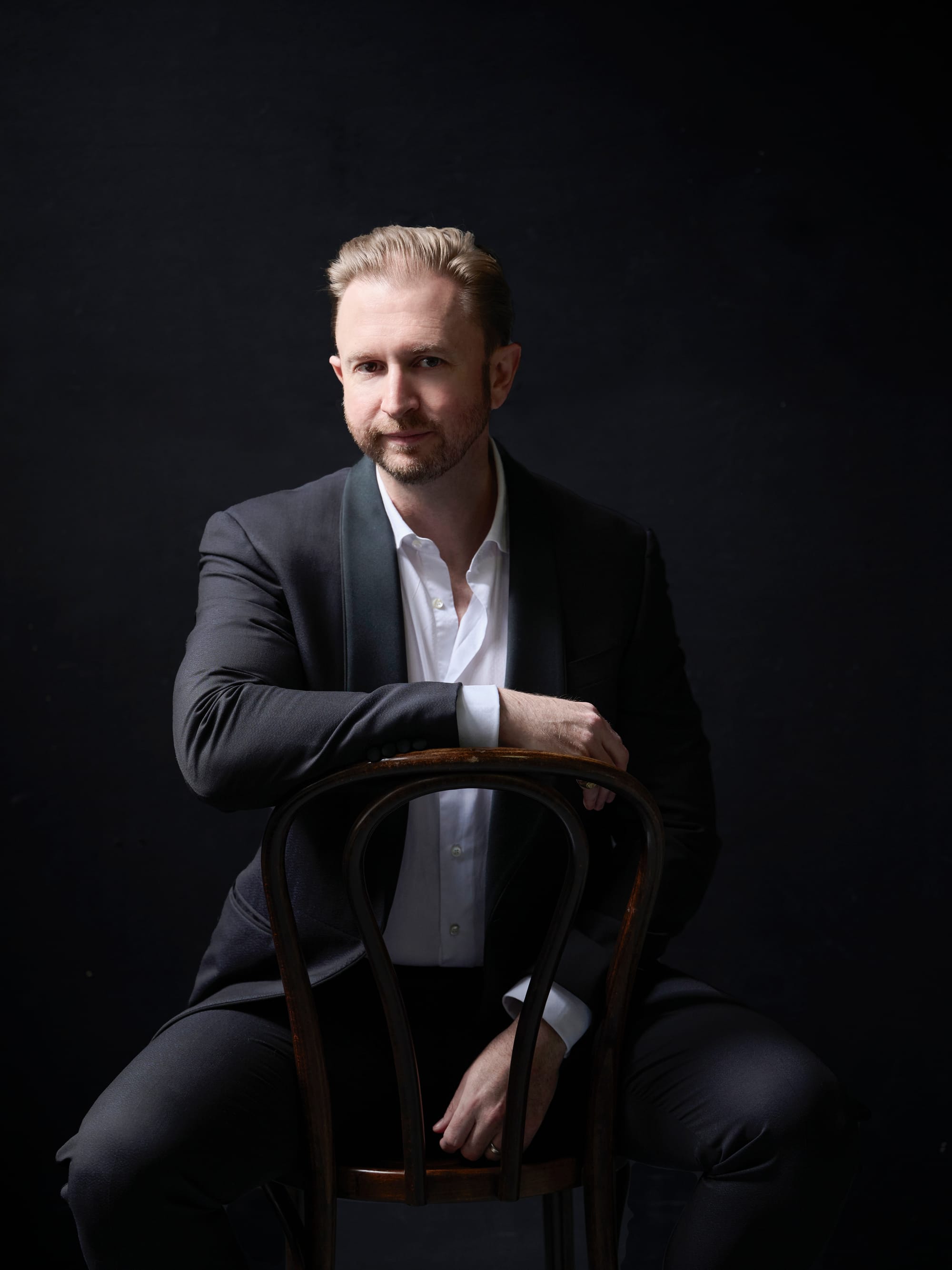 Robert Winter seated on a bentwood chair in a dinner jacket, against a dark background.