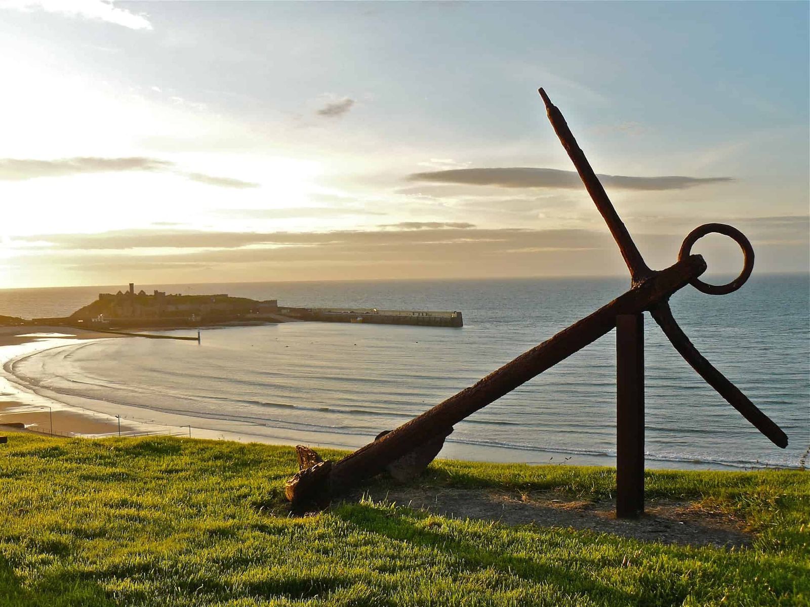 Photograph of an anchor resting on grass with Peel Beach and Peel Castle in the background.