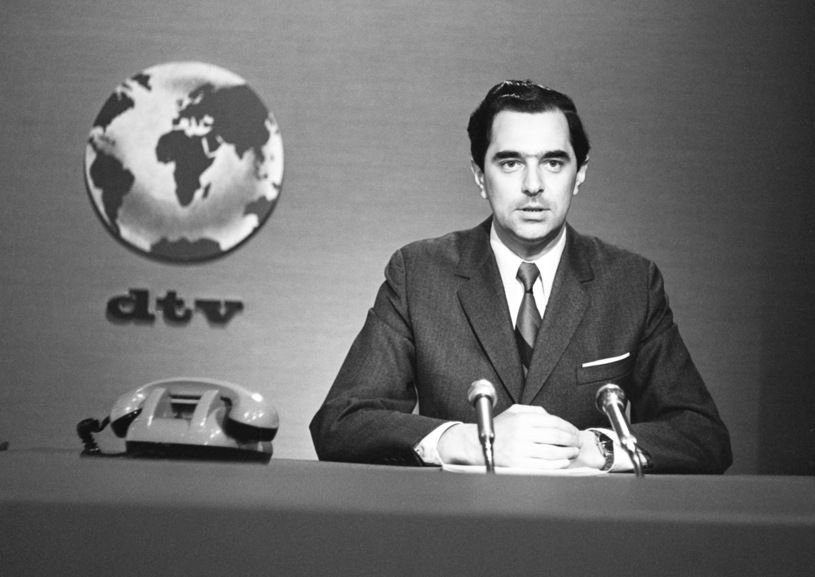 A black-and-white image of a serious-looking male news anchor (Andrzej Kozera) seated at a broadcast desk.