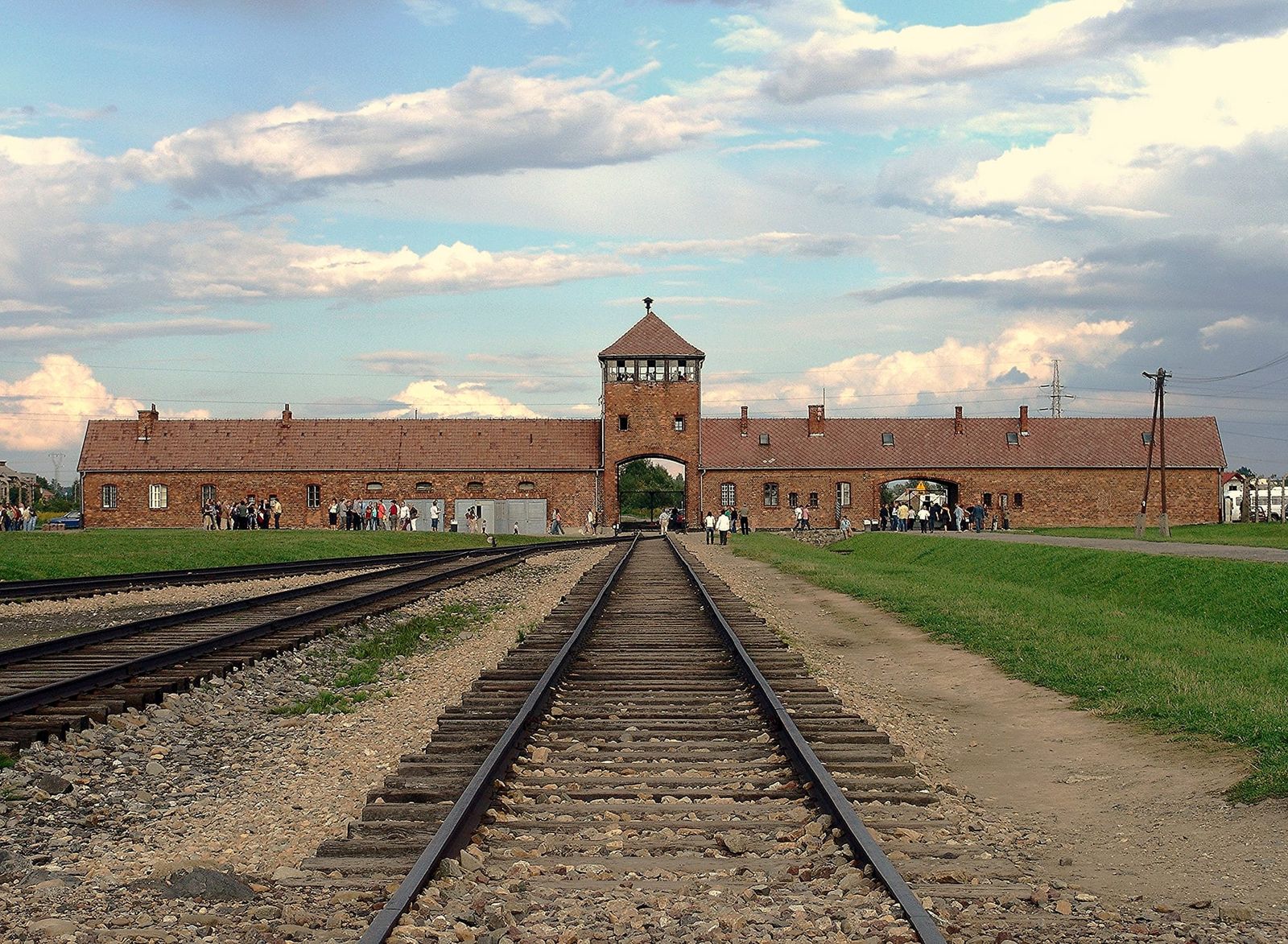 Railway tracks passing through the main gate of Auschwitz-Birkenau, with the brick watchtower and camp buildings visible beneath a wide sky, visitors near the entrance.