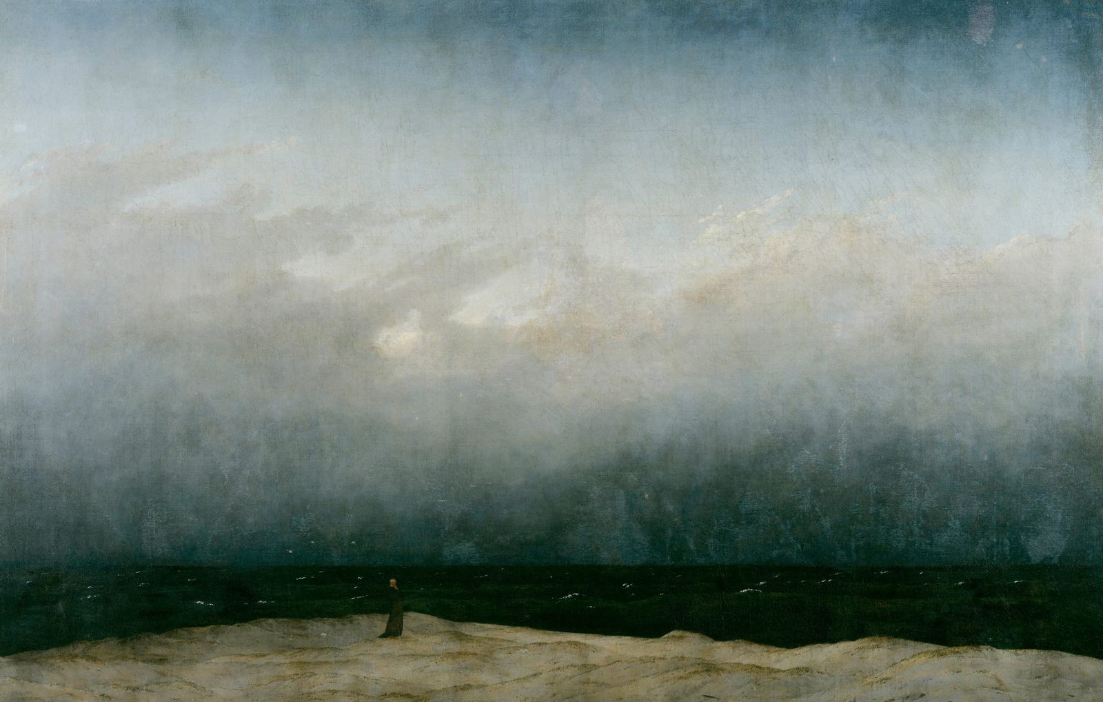 A lone monk stands on a dark shoreline beneath a vast, pale sky and empty sea; horizon blurred, land sparse, the small figure dwarfed by space and silence.