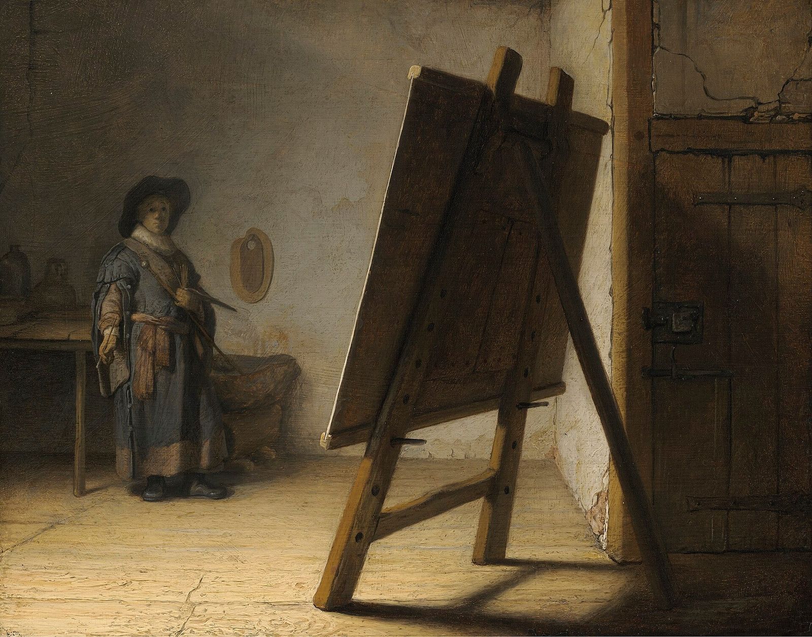 Rembrandt painting of a small figure in a tabard standing across a sparse studio, holding brushes and a palette, gazing at a large easel with its back turned to the viewer.