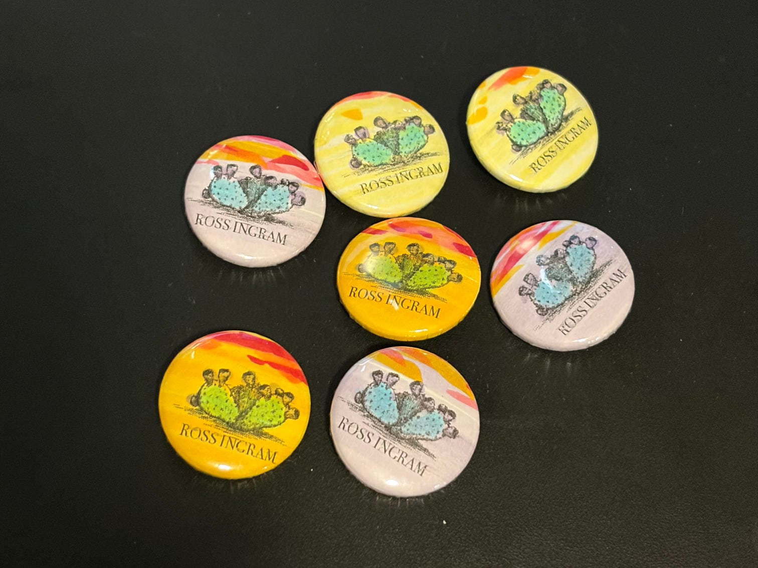 Handmade 1" pinback buttons featuring a print of a drawing of a prickly pear, each hand colored with alcohol markers.  Some have a yellow background, some orange, some lavender. The cactus is green or blue, with purple fruit.  All of the buttons feature a bright sunset sky.