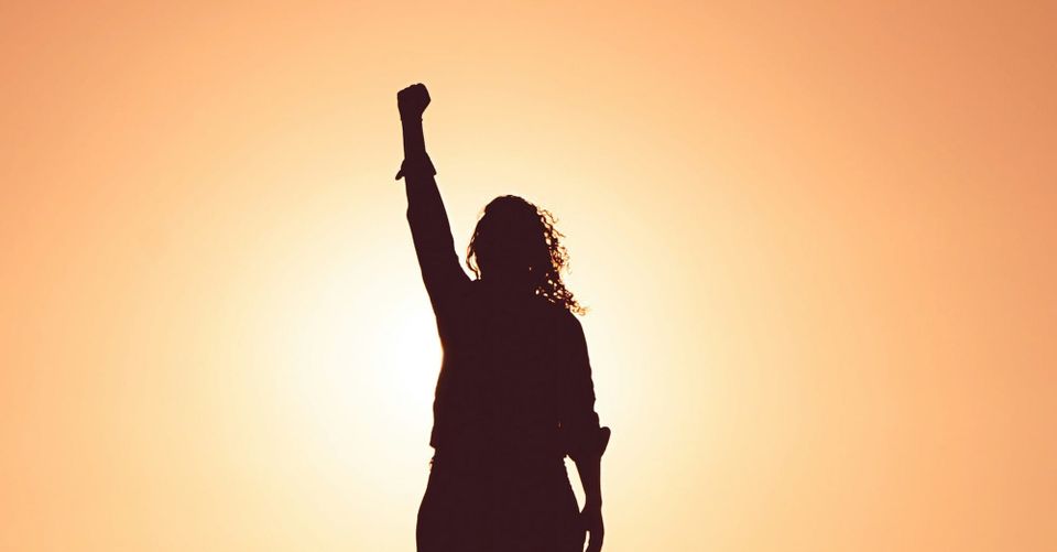 A silhouette of a person with long curly hair raises a fist in front of an orange sk