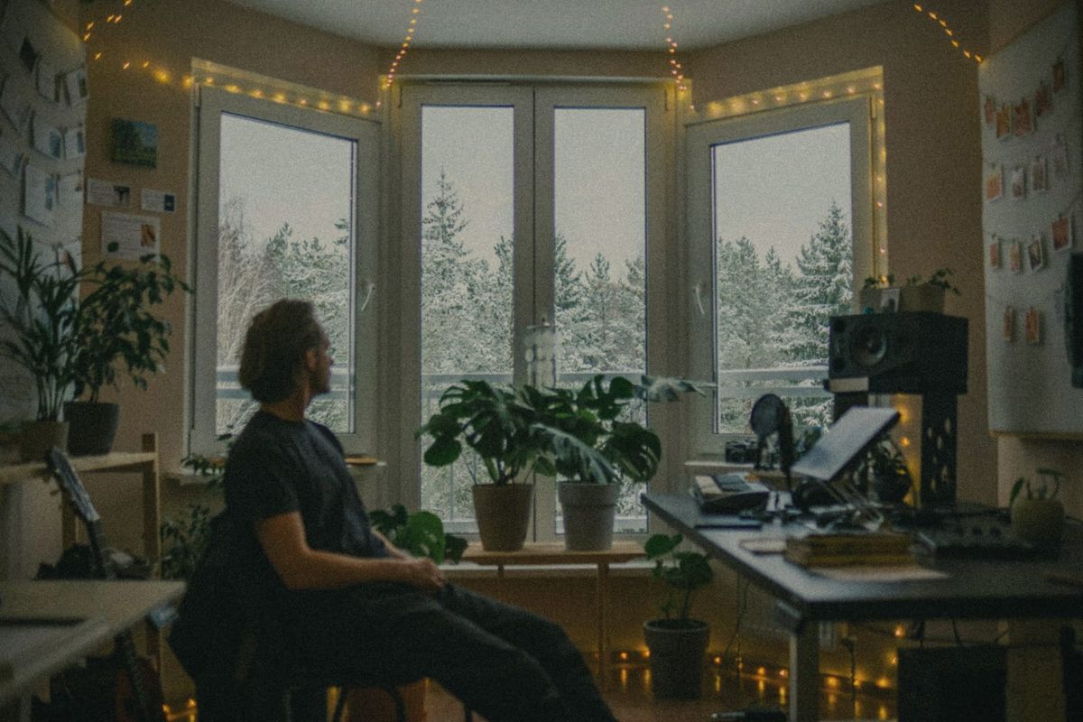 A man working at a desk in a warm, plant-filled home study with string lights, snowy trees beyond the window