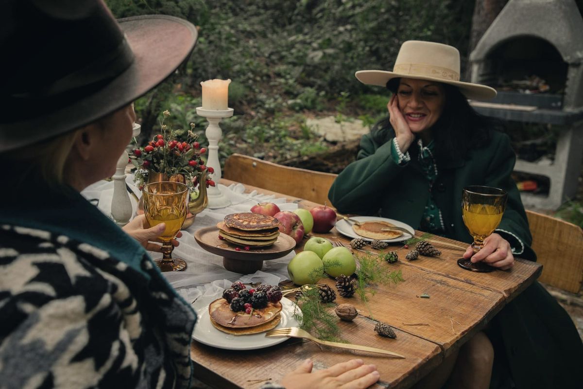 Two women sharing a laden autumn breakfast table outdoors, one smiling, warm candlelight