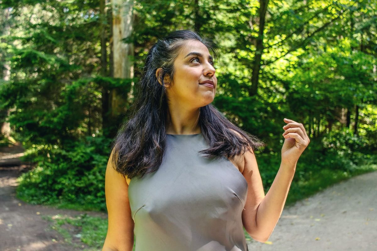 A woman looks up, smiling softly, on a sunlit green forest path in summer.