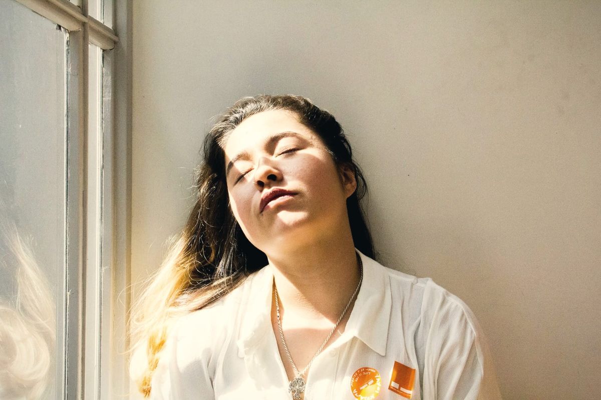 A woman by a sunlit window tilts her head back with her eyes closed, letting go.