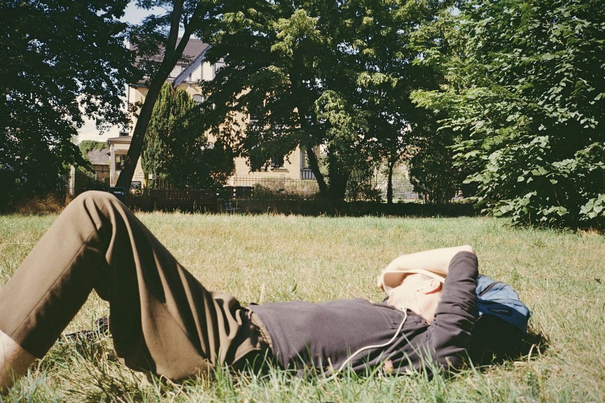 A man lying flat on a summer lawn in a park, one arm resting over his eyes